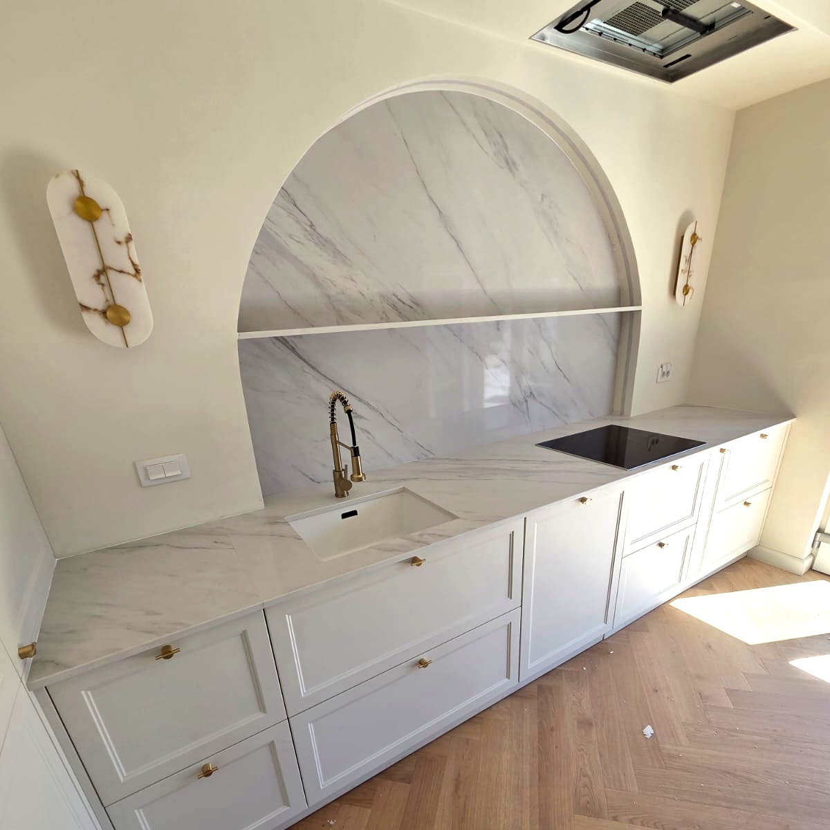 Vedere laterală bucătărie cu blat și panou arc din quartz Calacatta Riviera, mobilier alb cu mânere aurii — vinca.ro